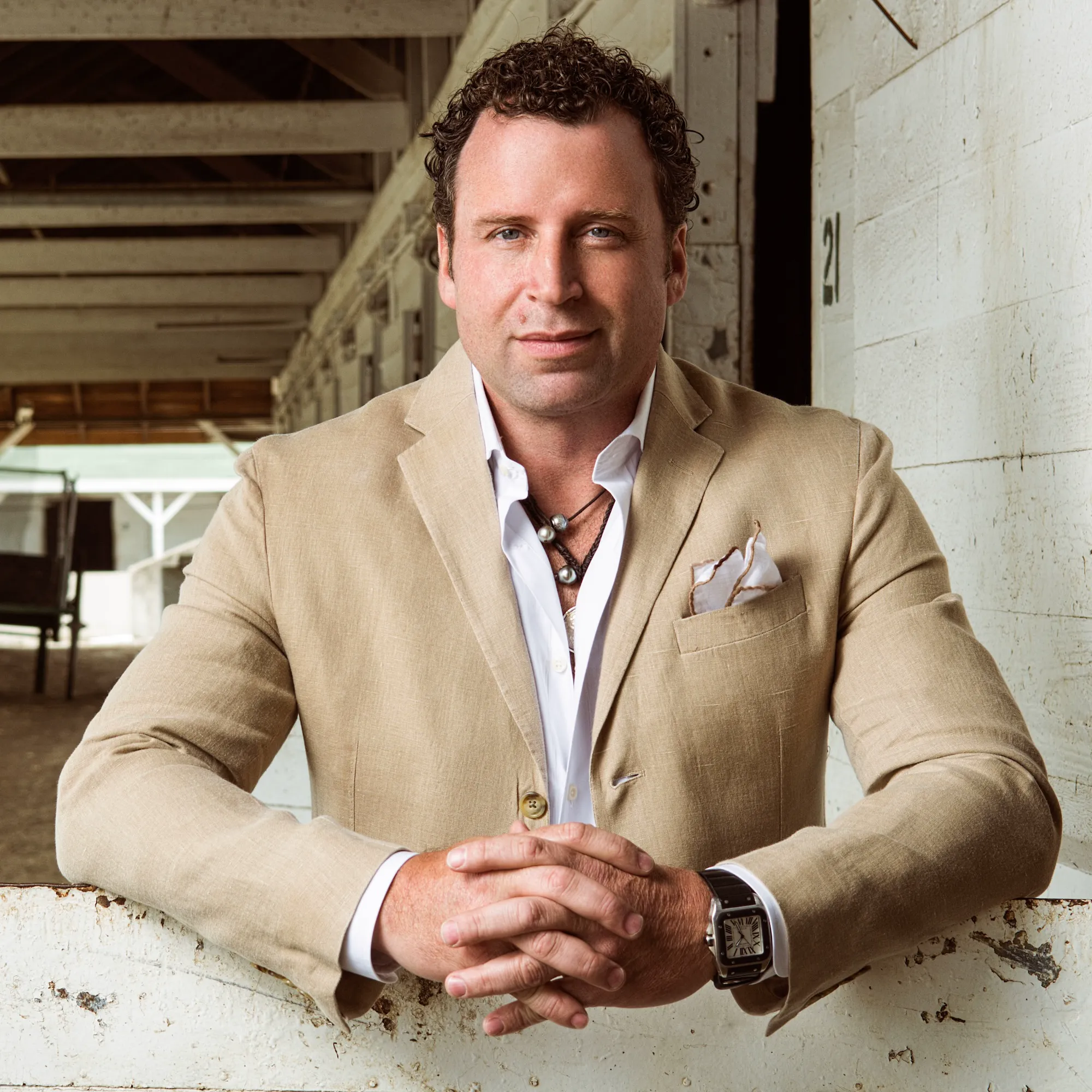 Vincent Peach, fine jewelry designer, in a tan jacket and white shirt, leaning on a weathered fence rail in a stable.